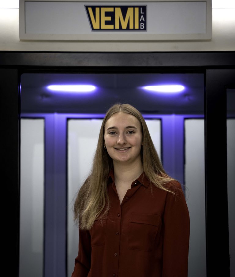 Rachel Coombs poses in front of the VEMI Lab sign.
